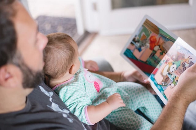 30 Must-Have Baby Books for Your Registry, a father sitting on the couch reading a book to his baby, who is leaning against him.
